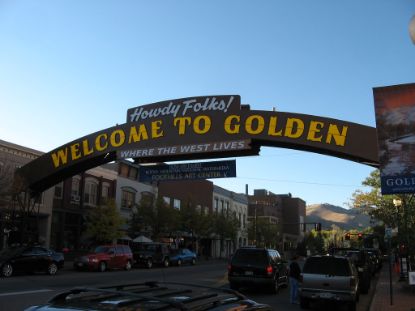 Street view of Downtown Golden featuring the large archway sign that reads “Howdy Folks! Welcome to Golden — Where the West Lives,” with shops, cars, and mountains in the background under a clear sky.